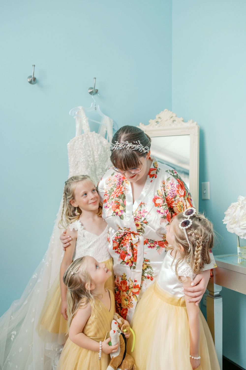 A woman in a floral robe shares a sweet moment with three young girls in tulle dresses in a light blue room.