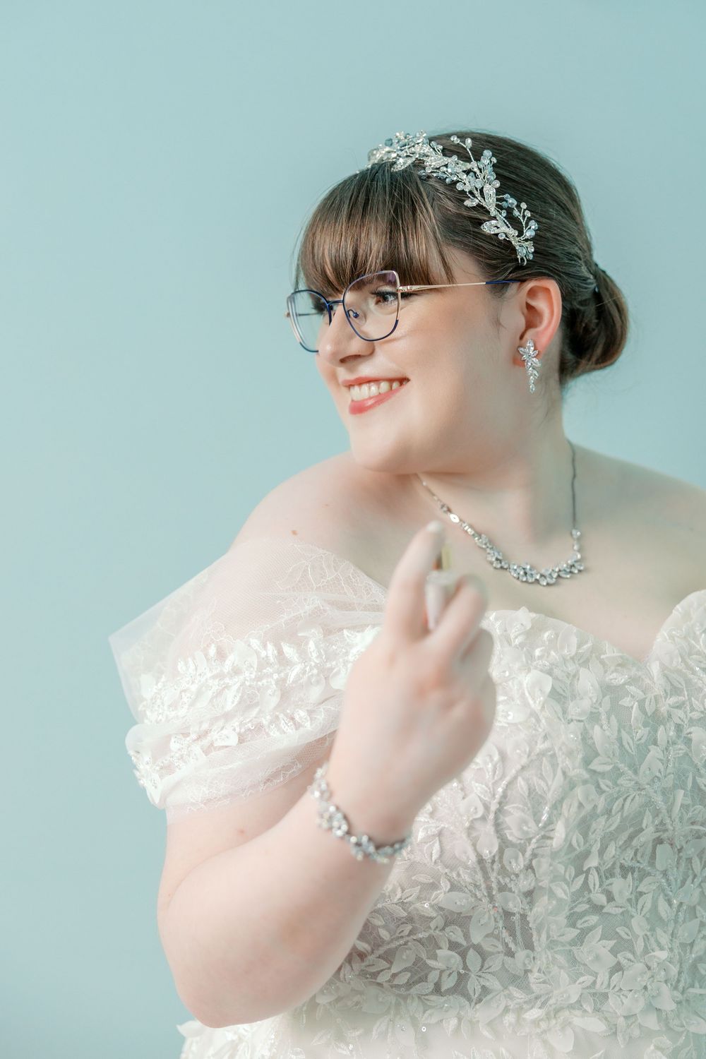 A bride in a white lace wedding dress wearing glasses, a crystal headpiece, matching necklace and bracelet against blue background.
