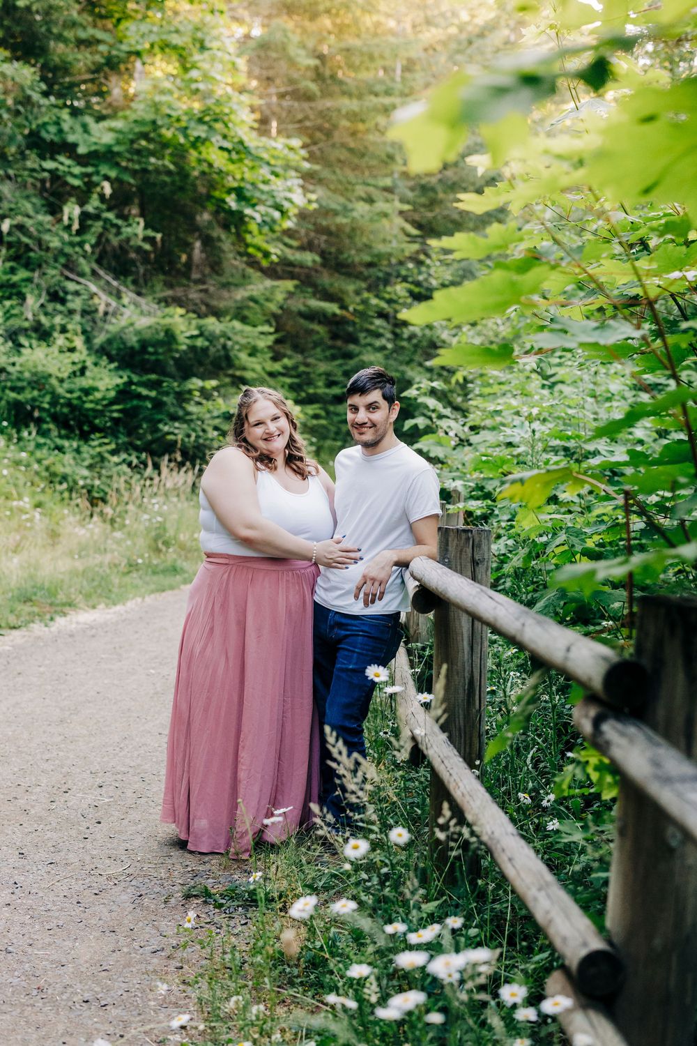 Couple in coordinated pink dress and white shirt pose together along a rustic wooden fence on a nature trail.