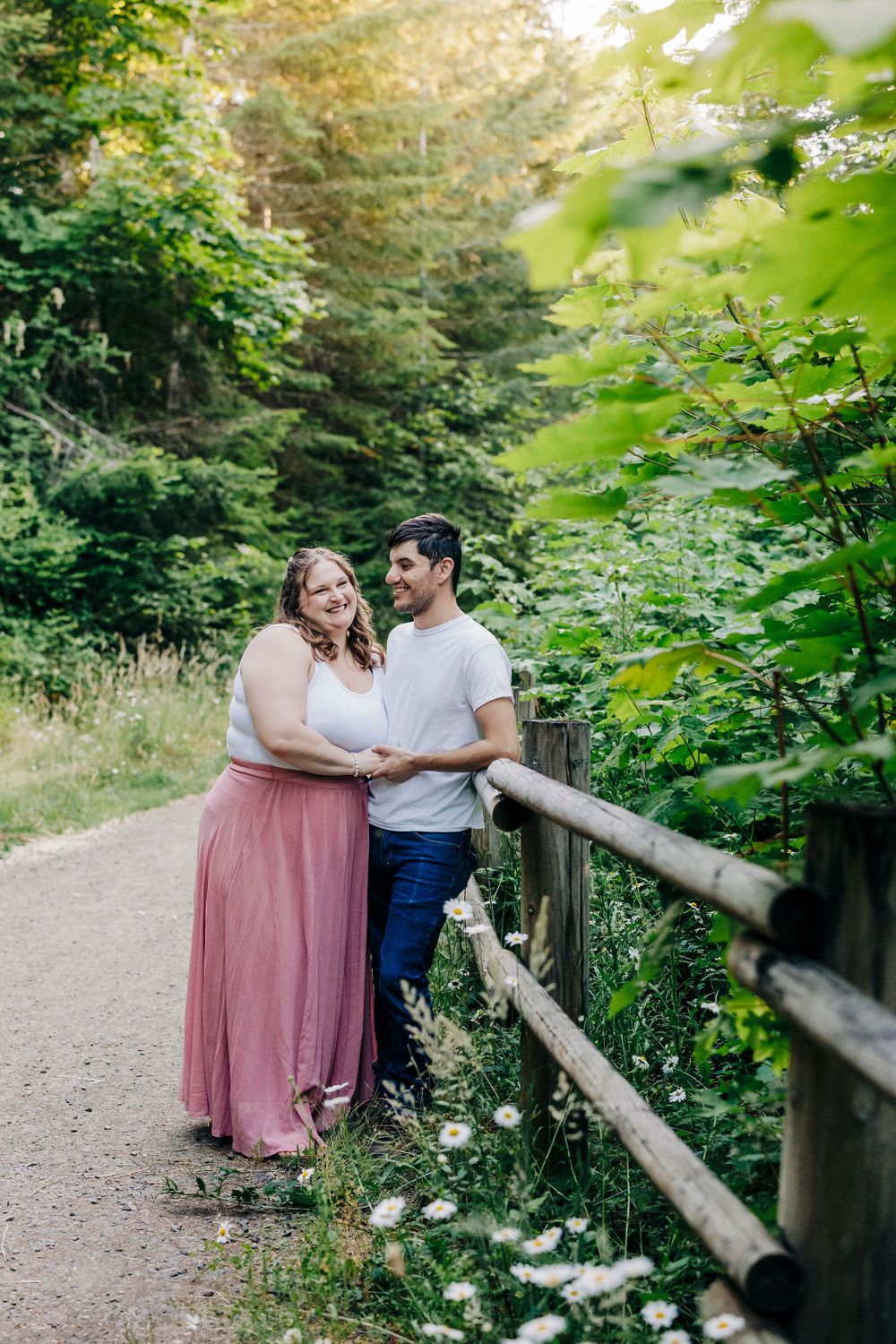 A romantic couple embraces on a tree-lined dirt path with rustic wooden railing and lush greenery.