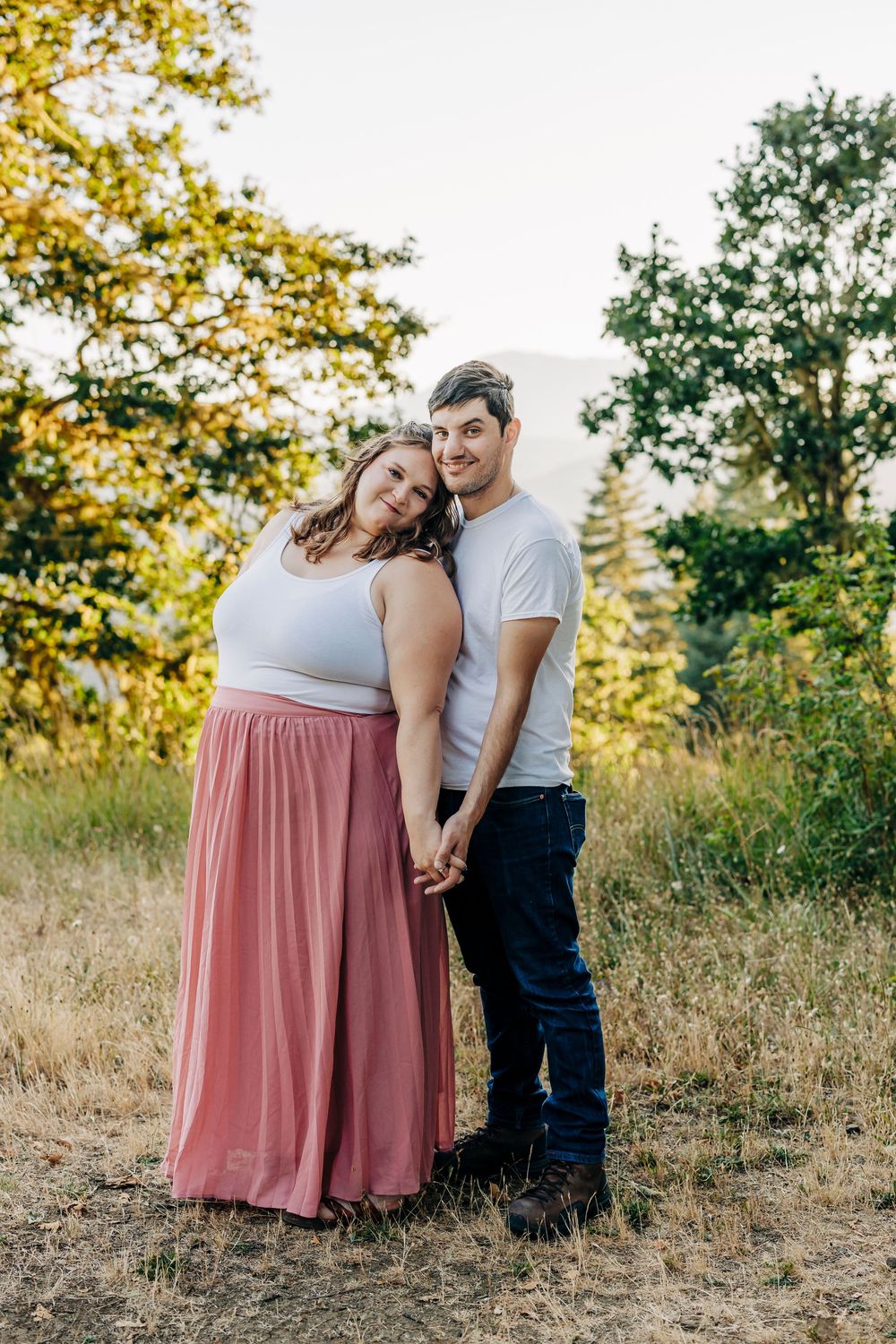 A romantic couple poses together in a sunlit meadow with yellow autumn trees, wearing casual attire and a flowing pink skirt.