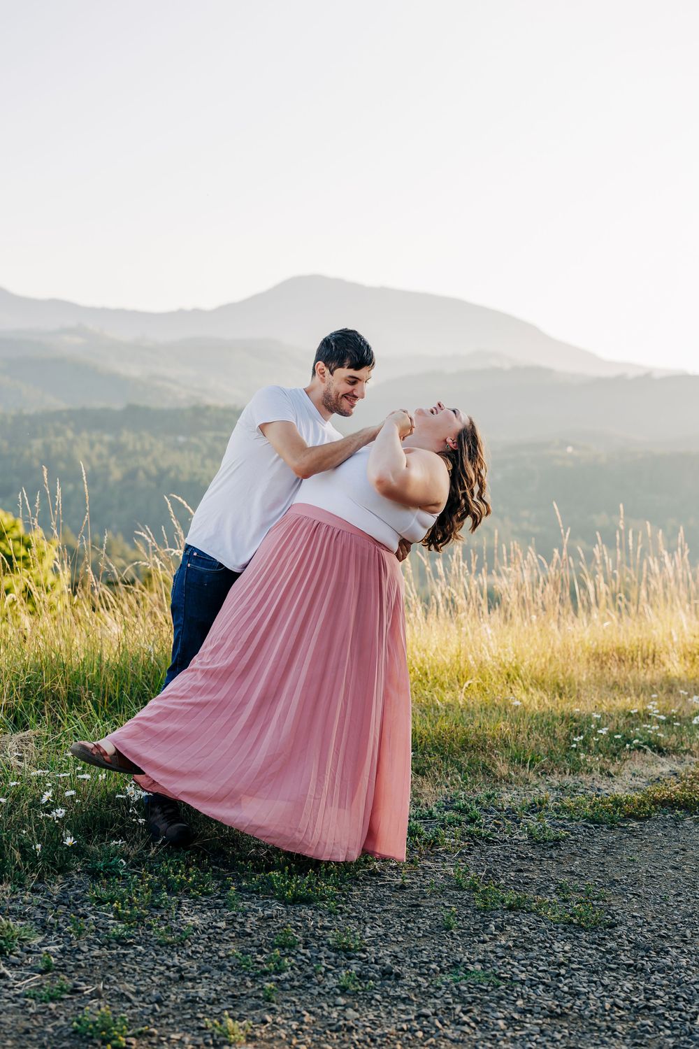 A couple shares a romantic moment against a mountain backdrop as the sunlight illuminates a golden field.