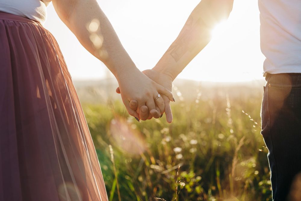 Two people holding hands in a sunny meadow at sunset with golden light filtering through tall grass and wildflowers.