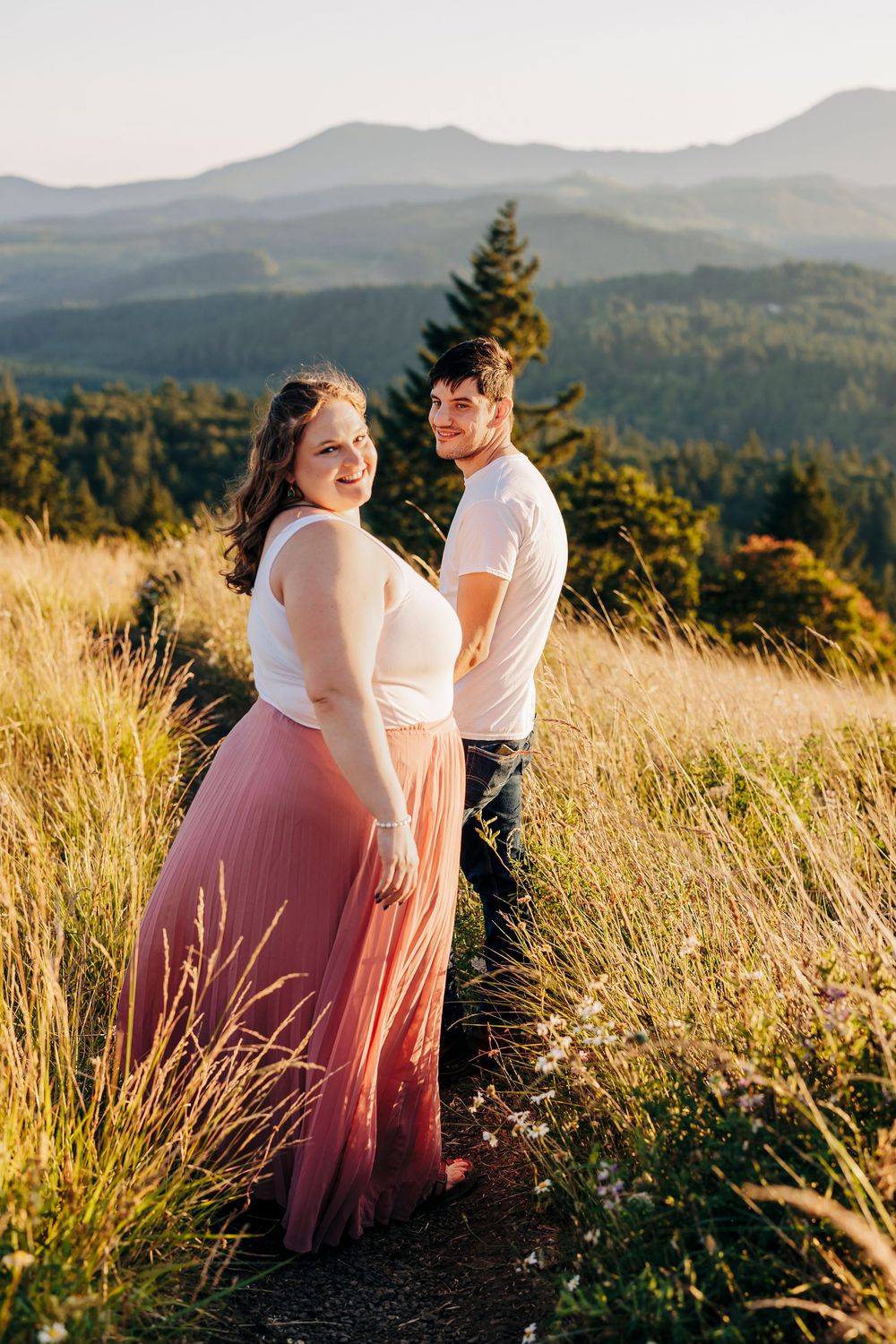 A romantic couple poses in golden sunlit meadow with mountains and evergreen forest in background during sunset.