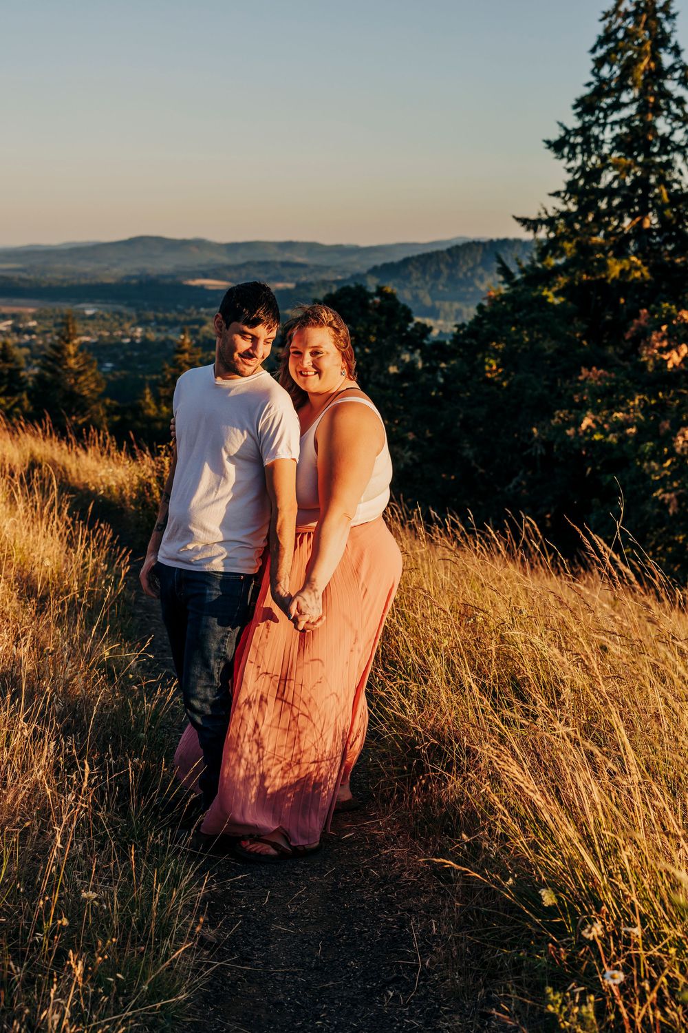 A couple embraces in a golden meadow during sunset with mountains and evergreen trees in the background.