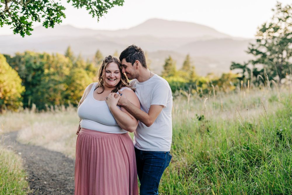 A couple embraces on a dirt path in a scenic mountain landscape with golden grass and trees at sunset.