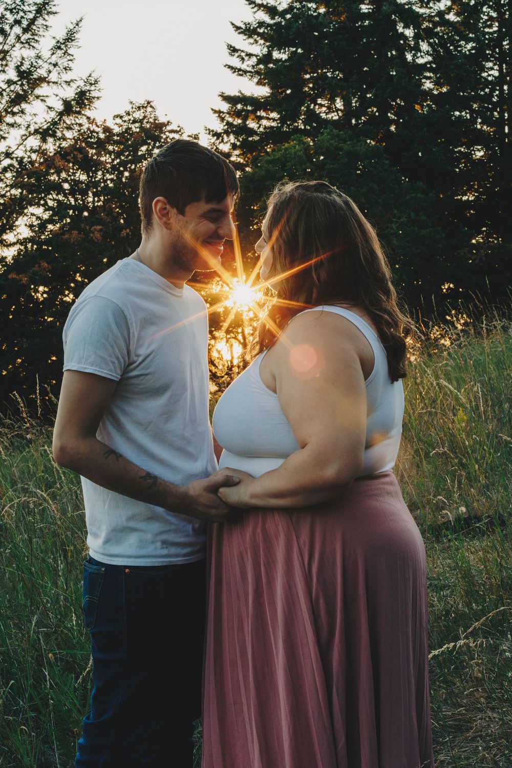 A couple embraces in a field at sunset with the sun's rays creating a starburst effect between them.