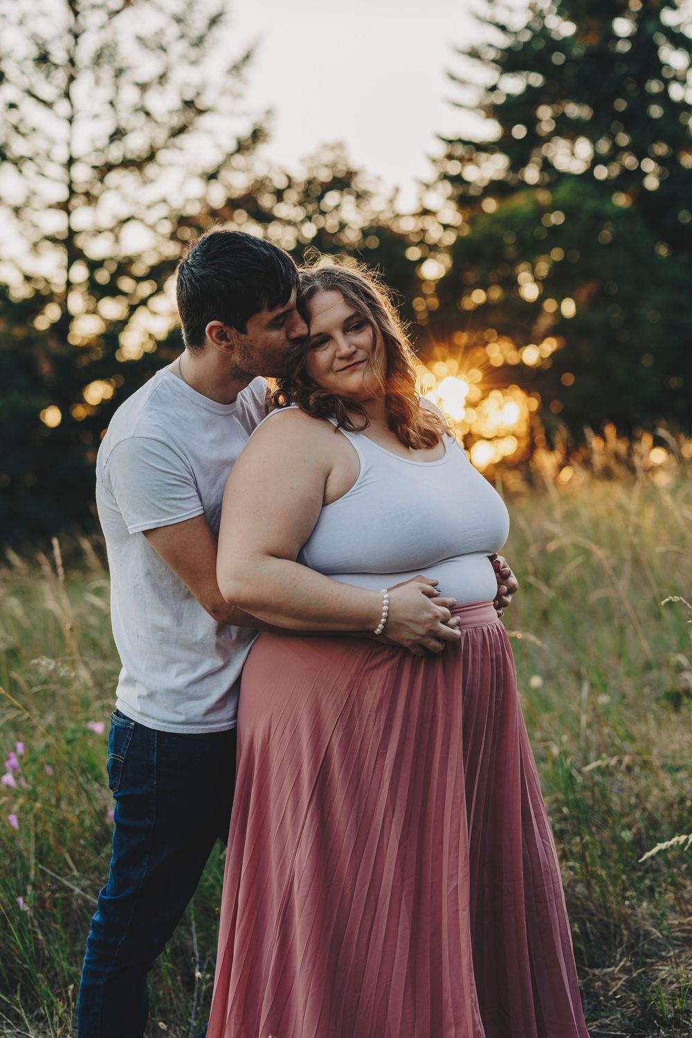 Romantic couple embraces in a sunlit meadow at golden hour, wearing casual clothes against a backdrop of evergreen trees.