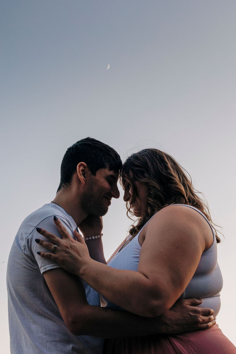 Intimate couple shares a tender moment against a dusky sky with a crescent moon visible in the background.