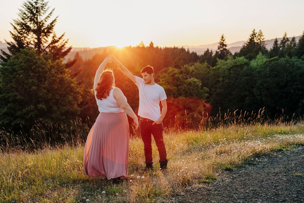 Silhouettes of couple standing on hilltop path with backlit golden sunset through evergreen trees in background.