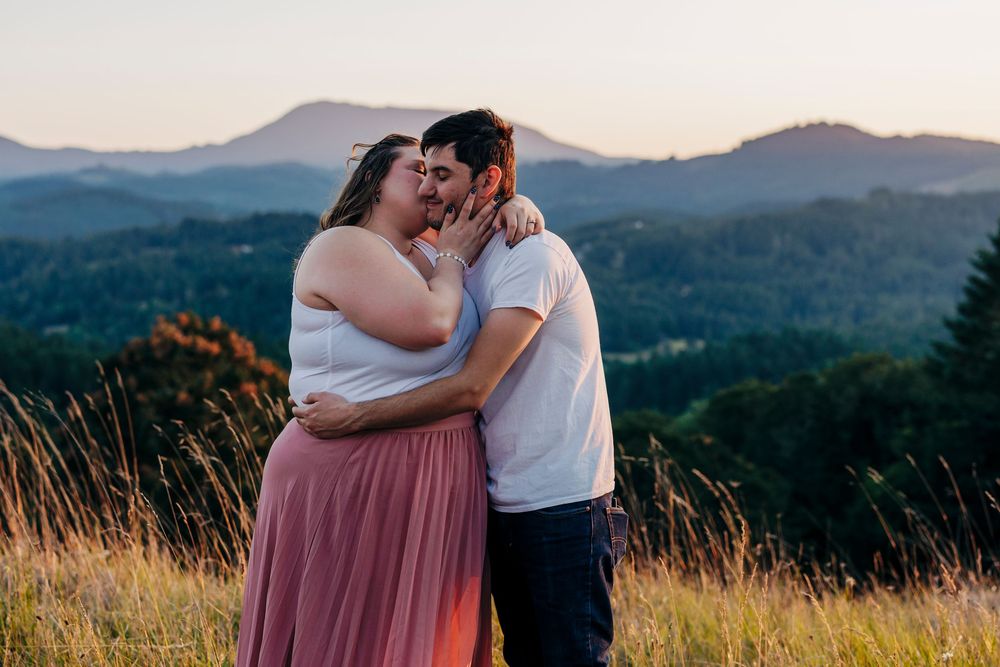 A couple embraces on a hilltop during sunset with mountains and forests in the background.