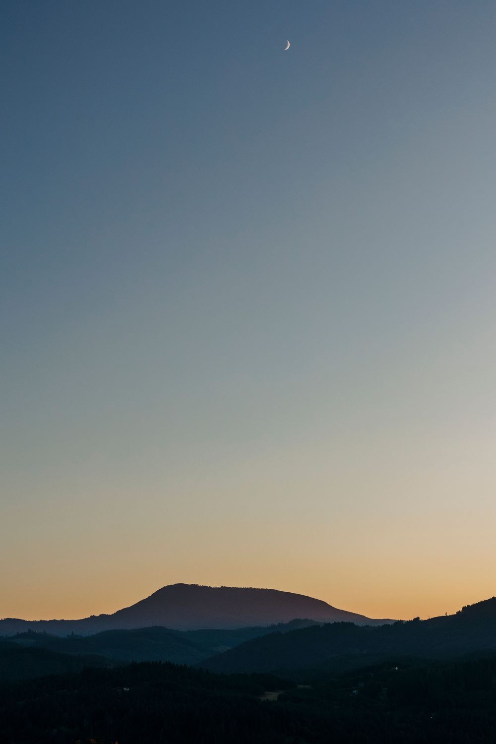 Crescent moon hangs in dusky blue sky above silhouetted mountain range at twilight.