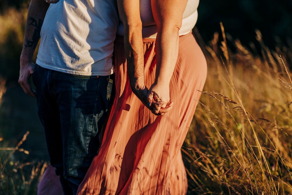 A couple stands hand-in-hand in a golden sunlit meadow wearing casual summer clothing.