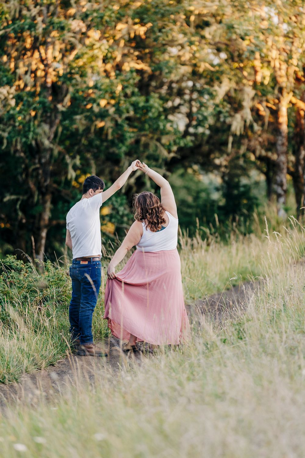 A couple twirls together in a sun-lit meadow with tall grass and trees creating a romantic summer scene.
