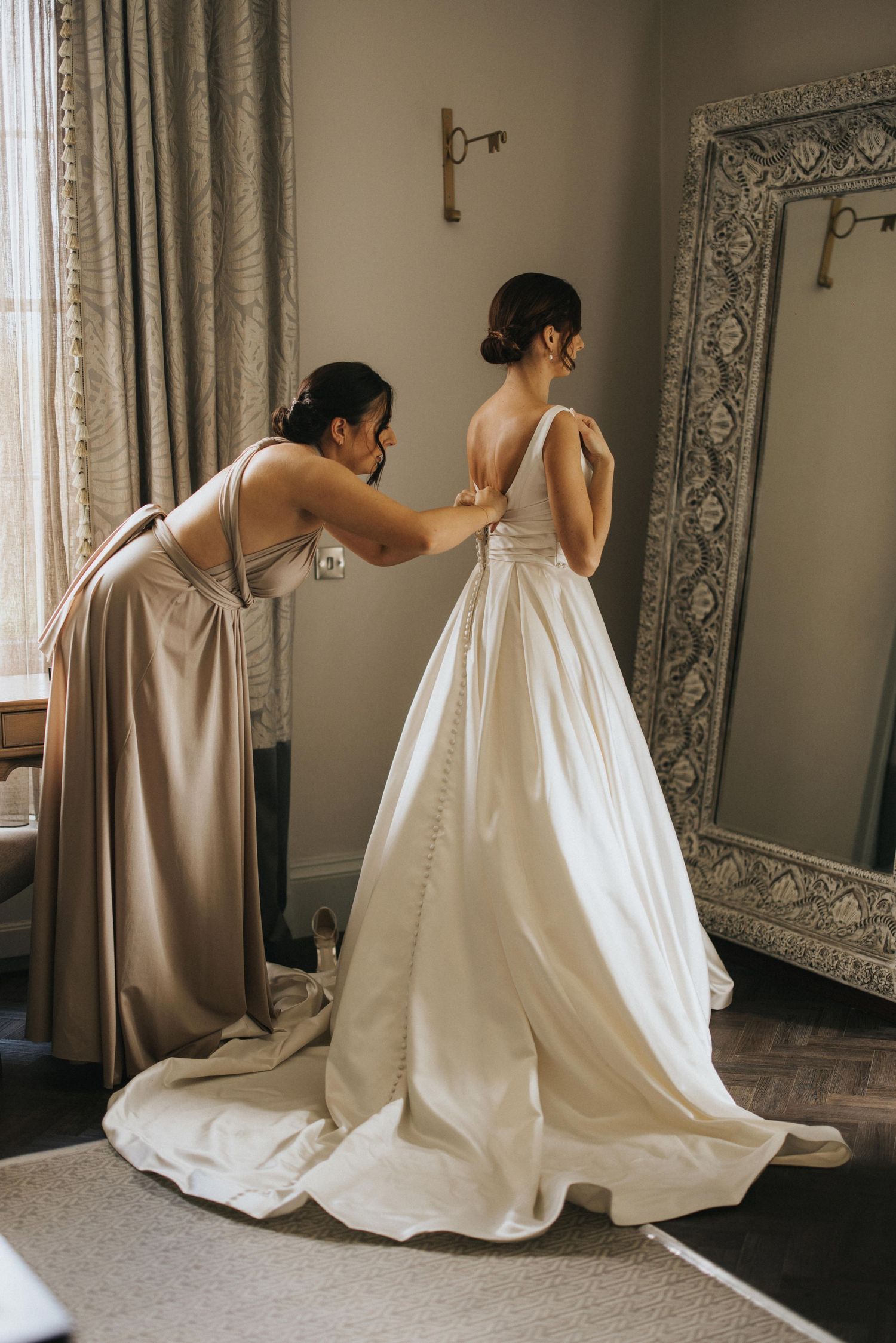 Bridesmaid adjusting a bride's white wedding dress while standing in front of an ornate mirror at Saltmarshe Hall.