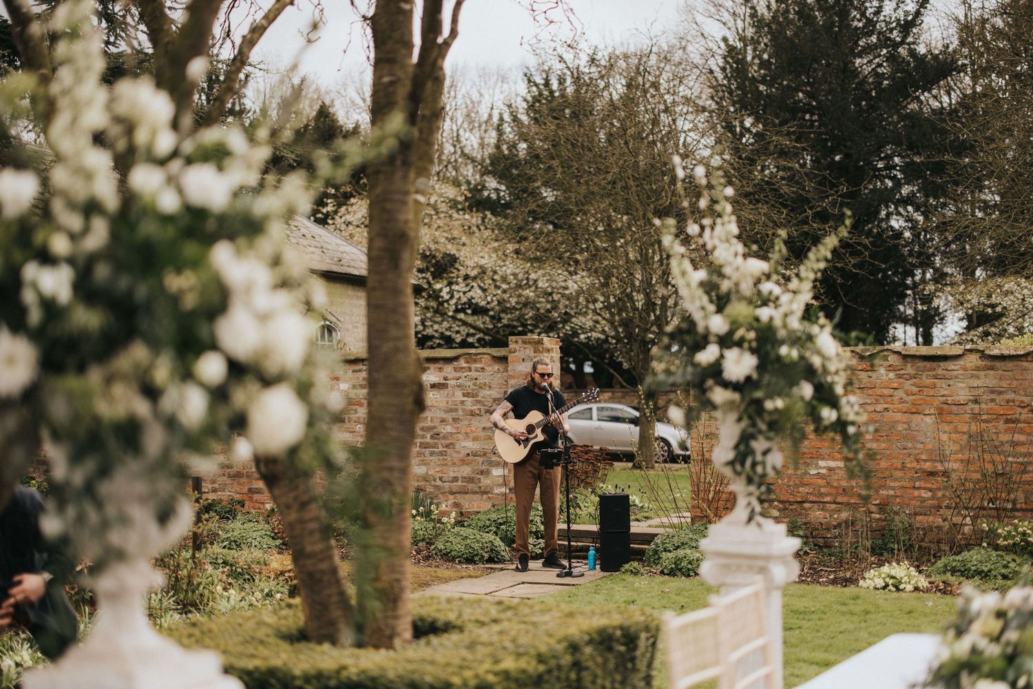 A live band plays music in a garden decorated with white floral arrangements near a brick wal at Saltmarshe Hall.