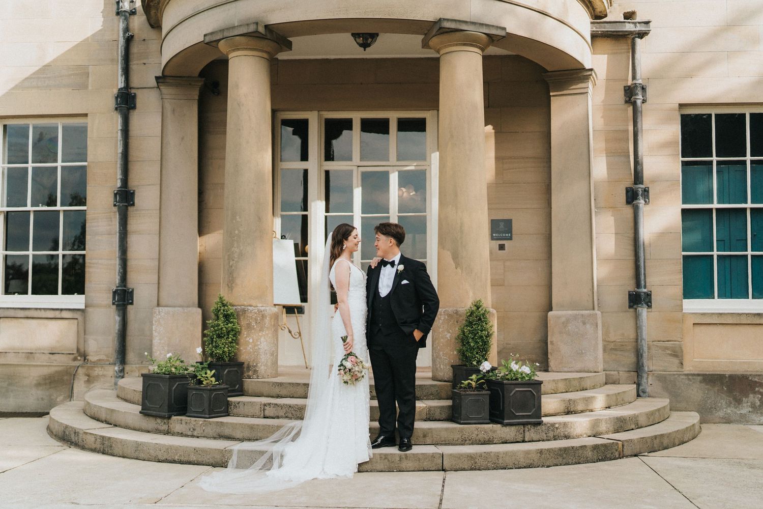 A couple poses on an elegant stone portico with columns and potted plants at Saltmarshe Hall entrance - taken by Grimsby photographer.