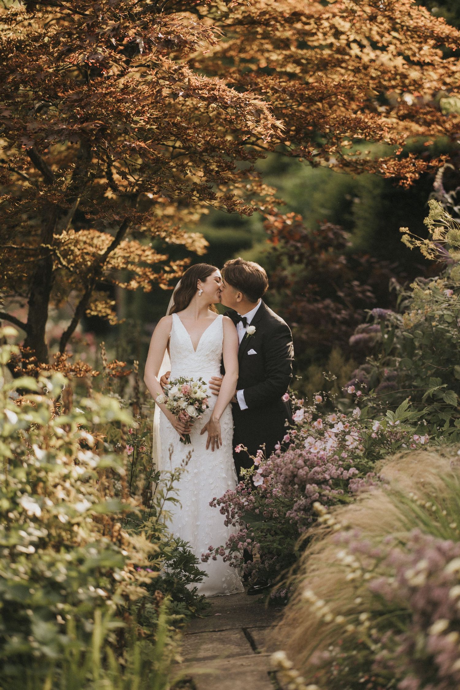 A couple stands intimately under autumn foliage on a garden path surrounded by blooming flowers at Saltmarshe Hall.