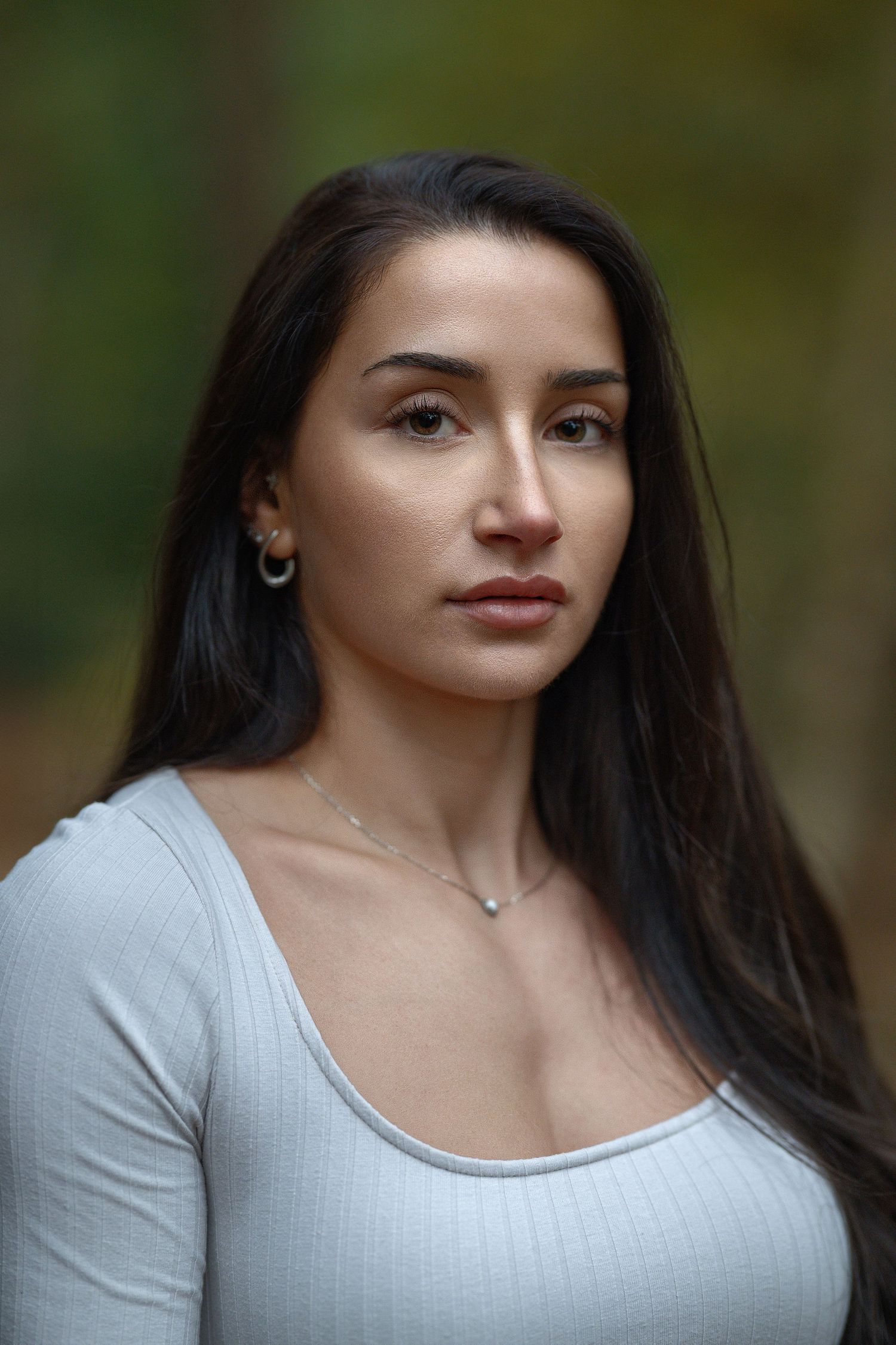 Portrait of a person with long dark hair wearing a white scoop neck top against a blurred green background.