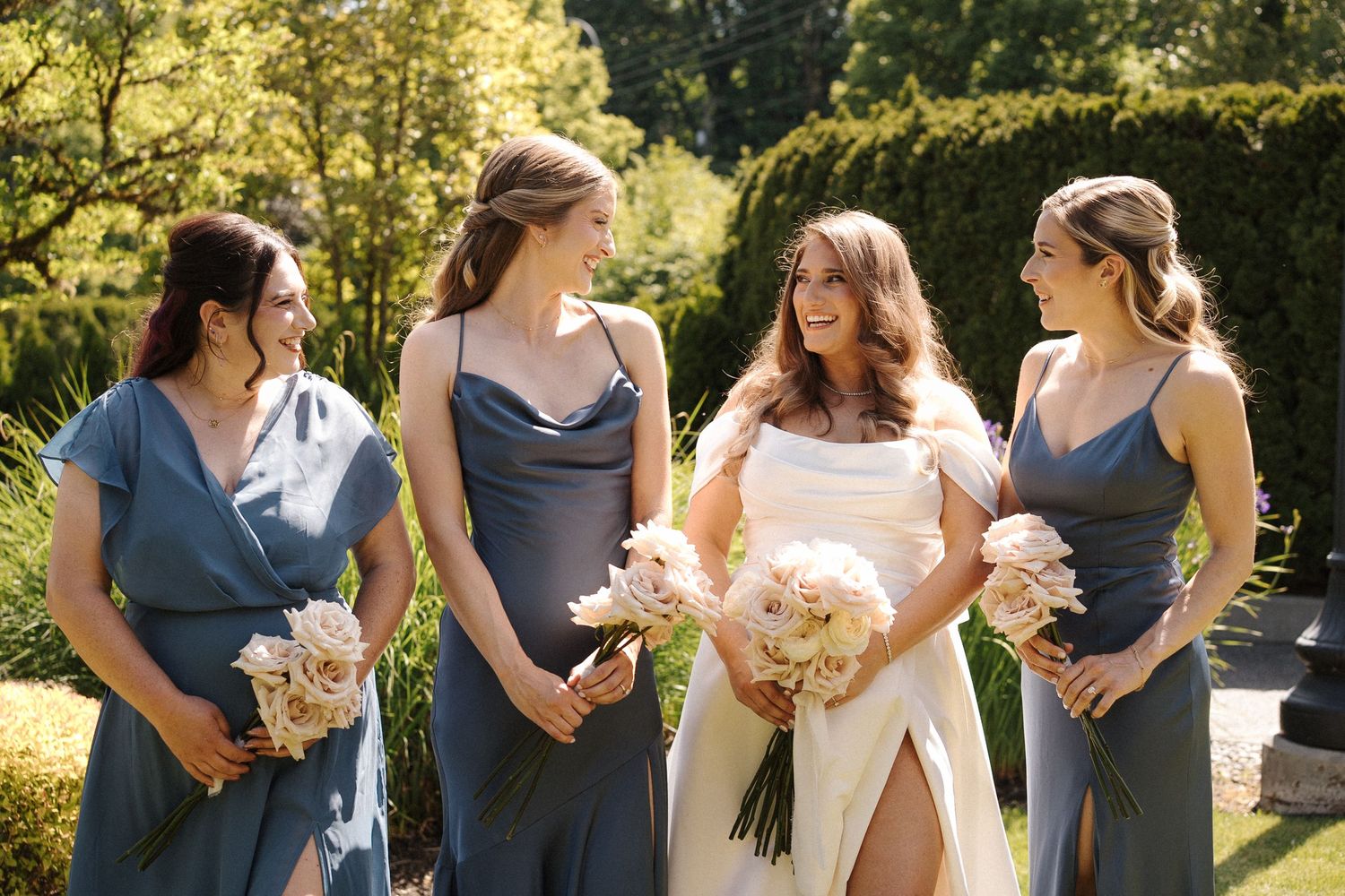 A wedding party wearing blue bridesmaid dresses and carrying pink floral bouquets stands together outdoors.