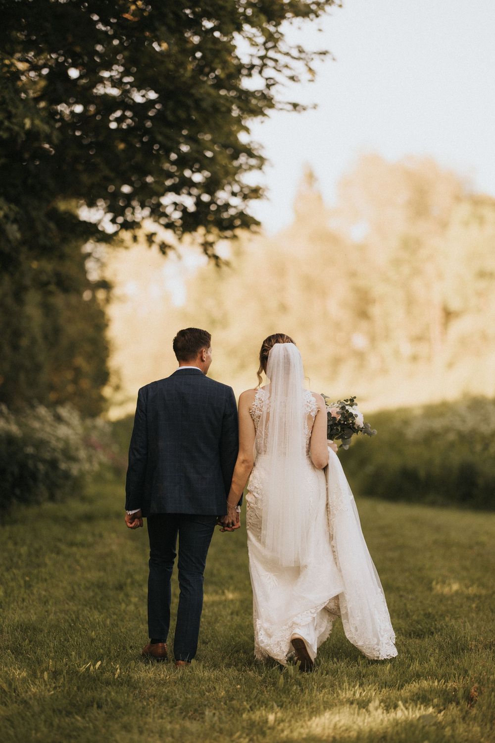 A newlywed couple walks hand in hand through a sunlit meadow on their wedding day at Hall Farm - image captured by Grimsby photographers.