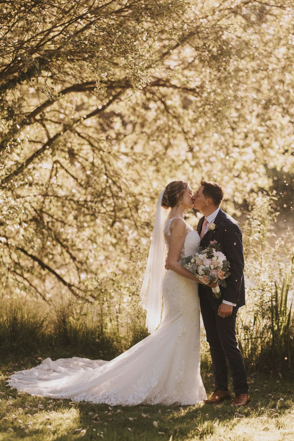 Couple shares romantic moment under sunlit trees with bride wearing flowing white gown at Hall Farm - image captured by Grimsby photographer
