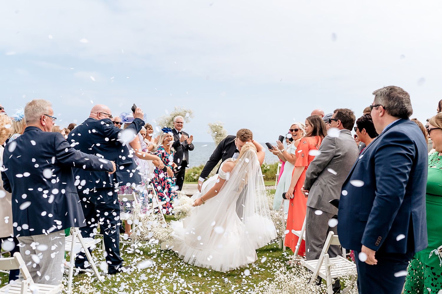 Wedding guests shower white confetti over a couple during an outdoor celebration on a bright sunny day.