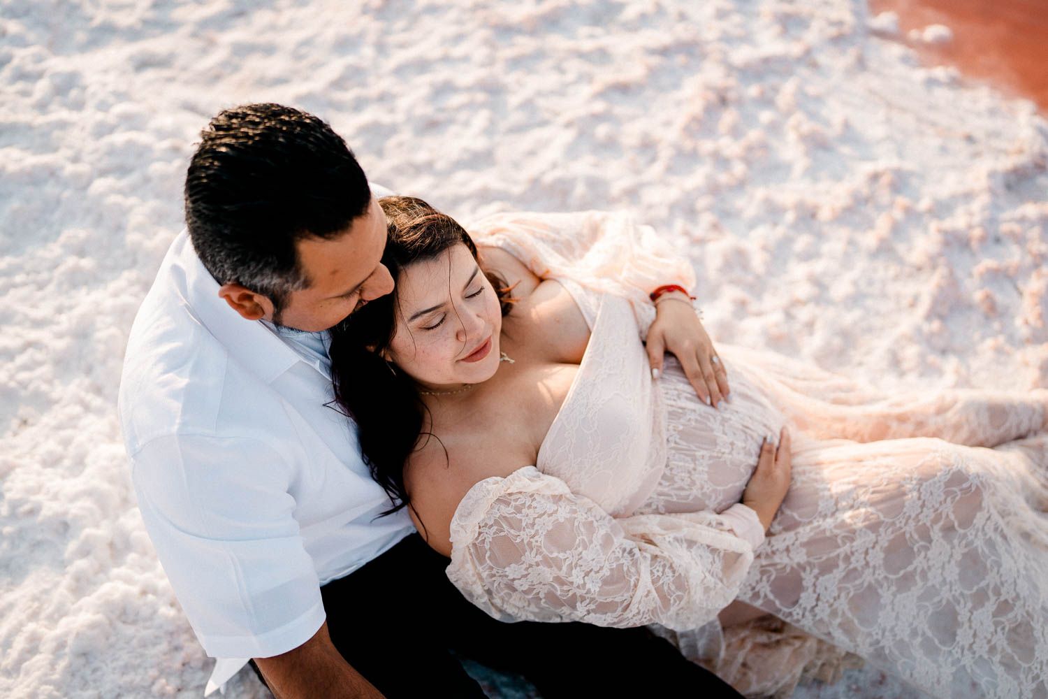 A romantic couple in white attire embraces on sandy beach at golden hour.