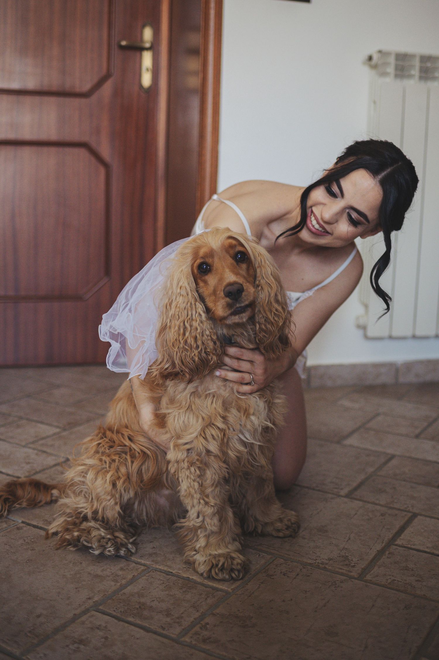 A person in white embraces a golden Cocker Spaniel dog while sitting on tiled floor near a wooden door.