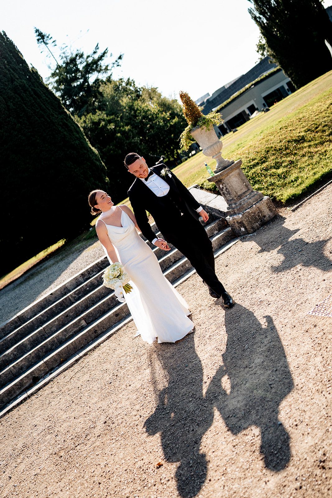 A couple in wedding attire being photographed on stone steps, casting long shadows on a sunny afternoon.