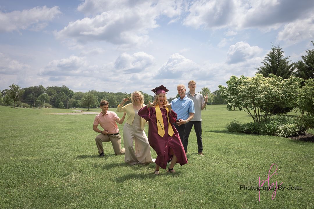 People celebrating graduation with playful poses on a sunny day in a lush green field with cloudy sky backdrop.