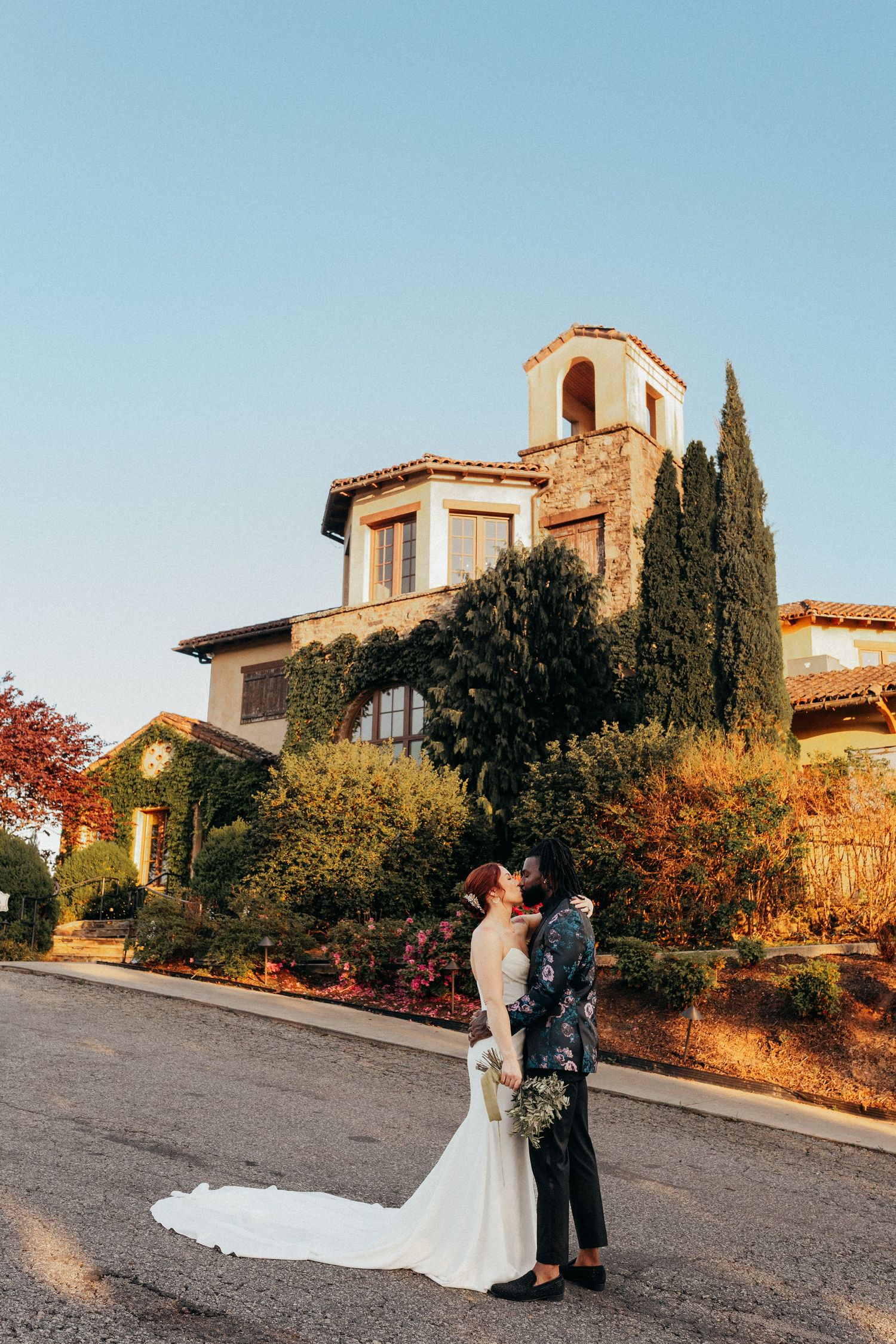 Two people share an intimate moment at sunset outside a Tuscan-style villa with cypress trees and autumn foliage.