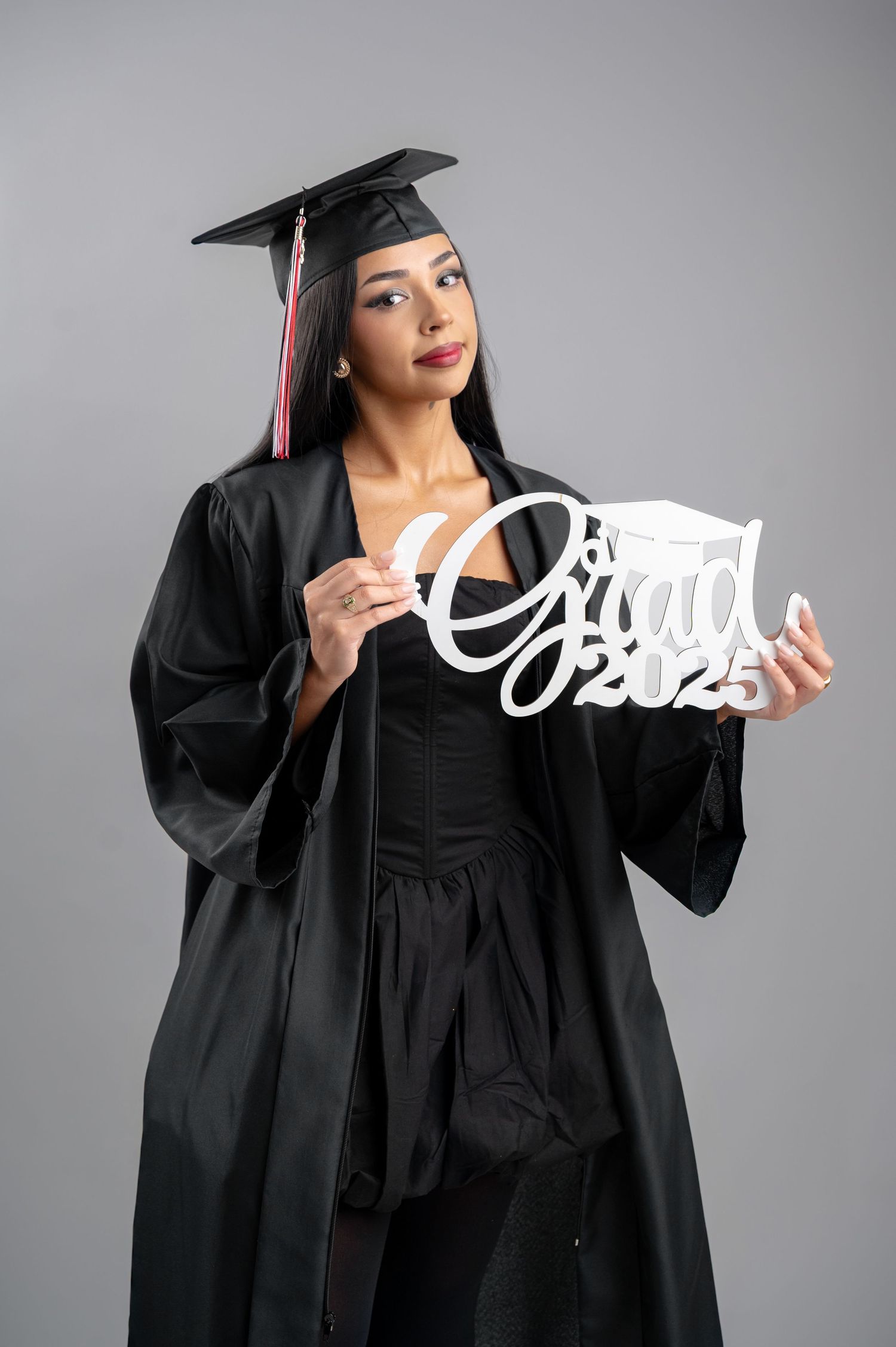 Graduate in black cap and gown holding decorative Grad 2025 sign against gray background.