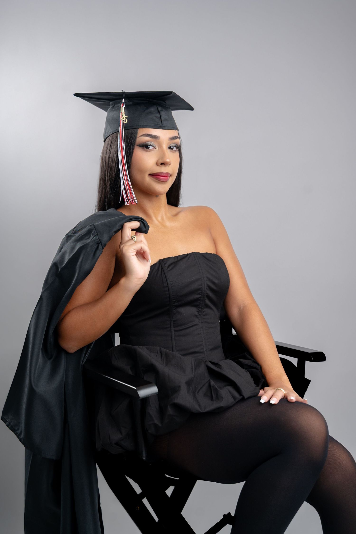 Graduate poses elegantly in black gown and cap on director's chair against grey backdrop.