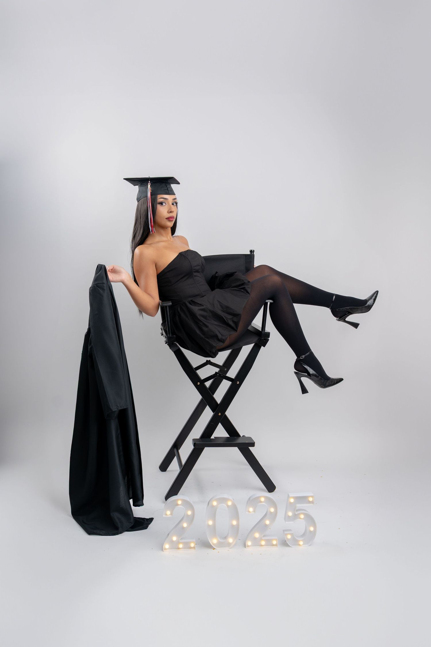Graduate in black dress and cap posing on director's chair with illuminated 2025 numbers below.