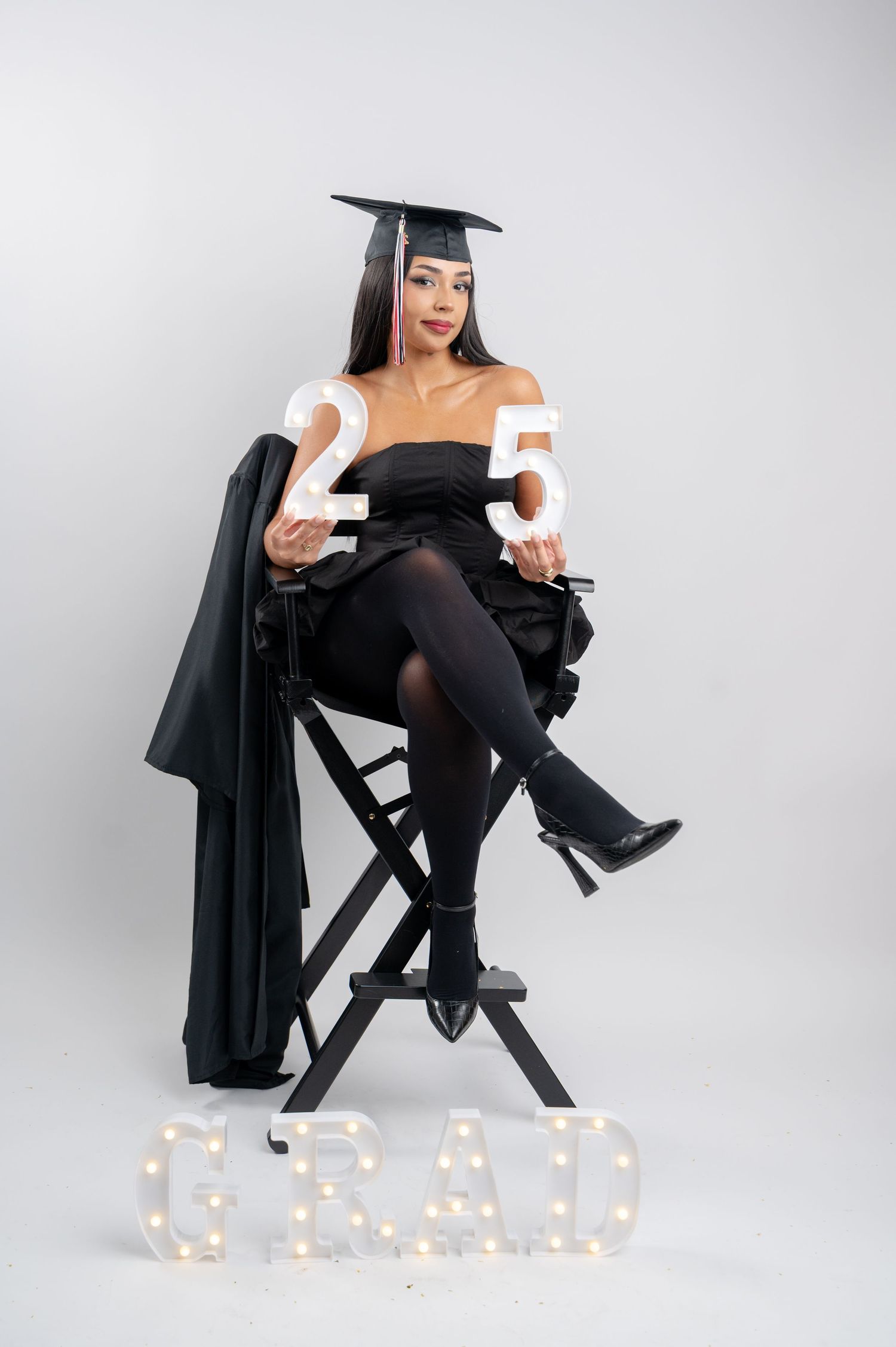 A graduation portrait featuring someone seated on a director's chair in black attire with illuminated GRAD letters below.