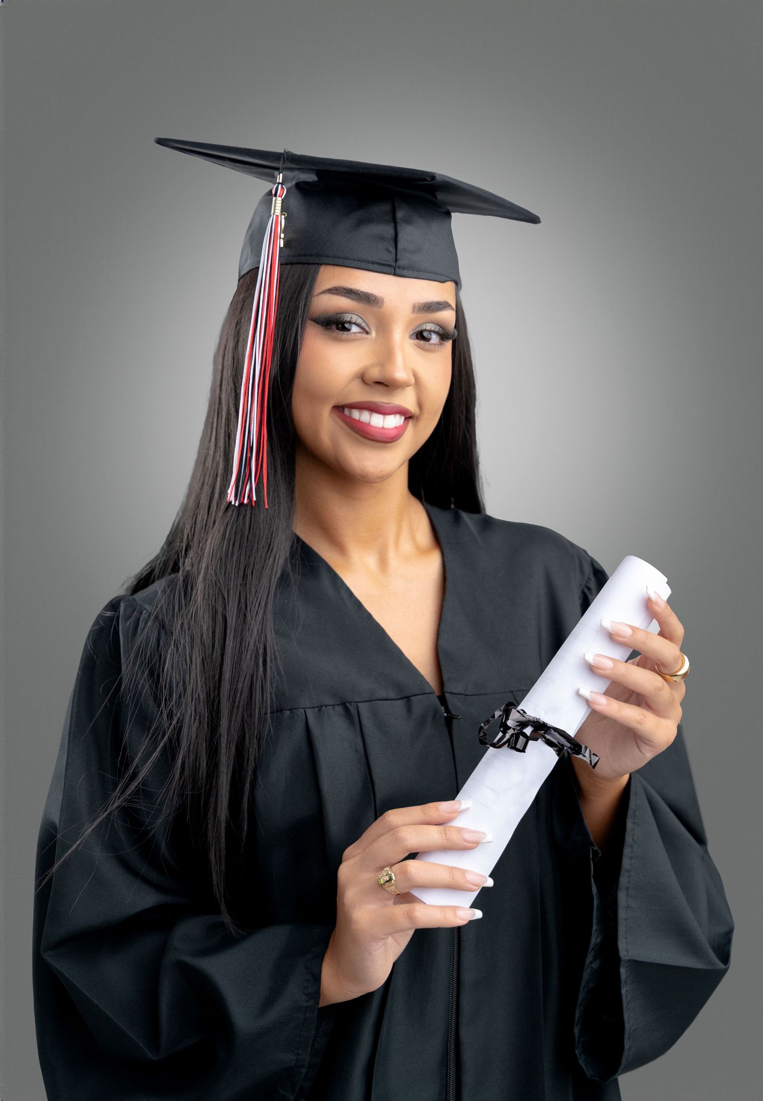 Graduate in traditional black cap and gown holds diploma while posing against gray background for graduation portrait.
