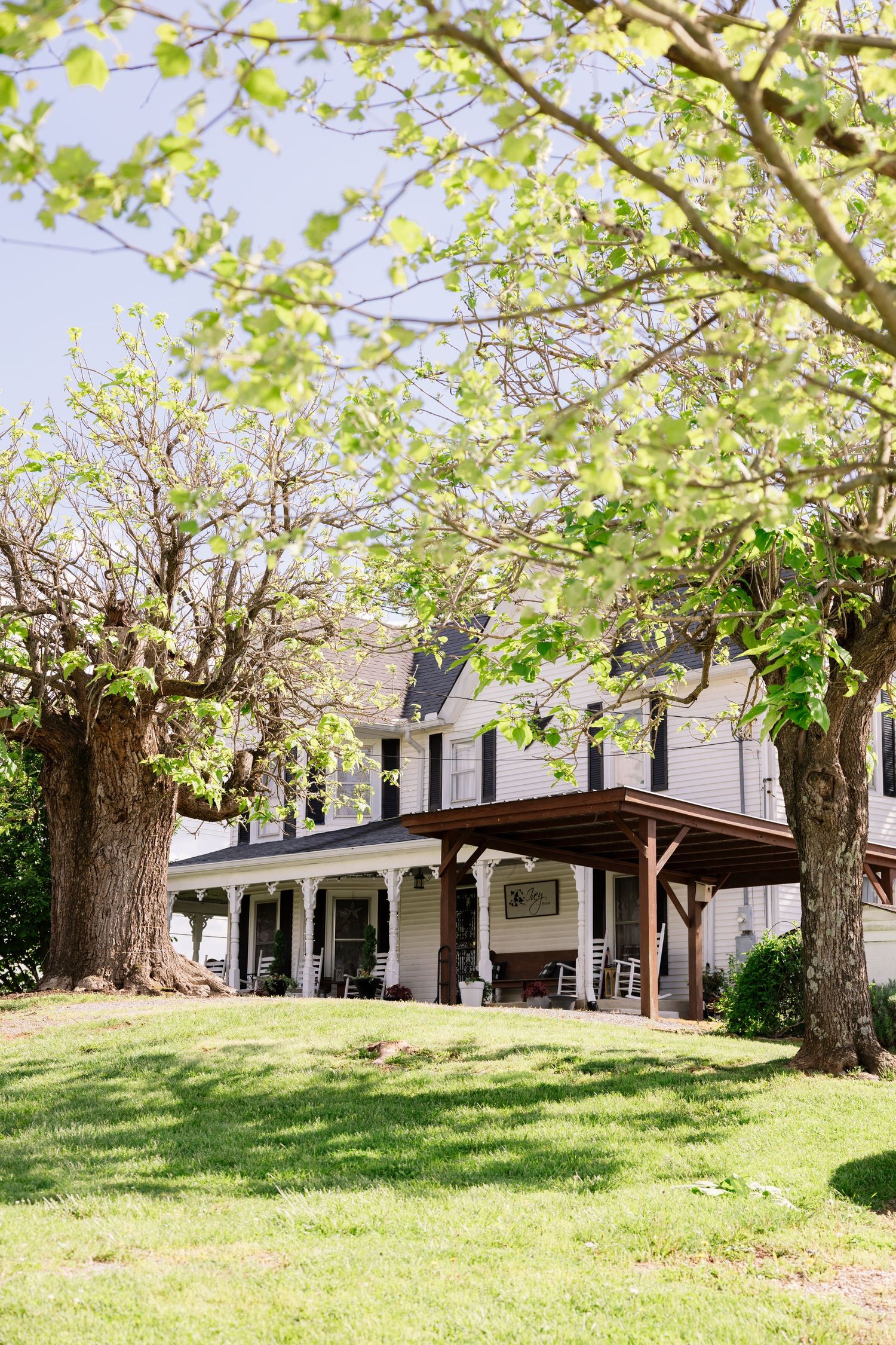 White farmhouse-style house with dormers and a wrap-around porch nestled among trees on a grassy hill in springtime.