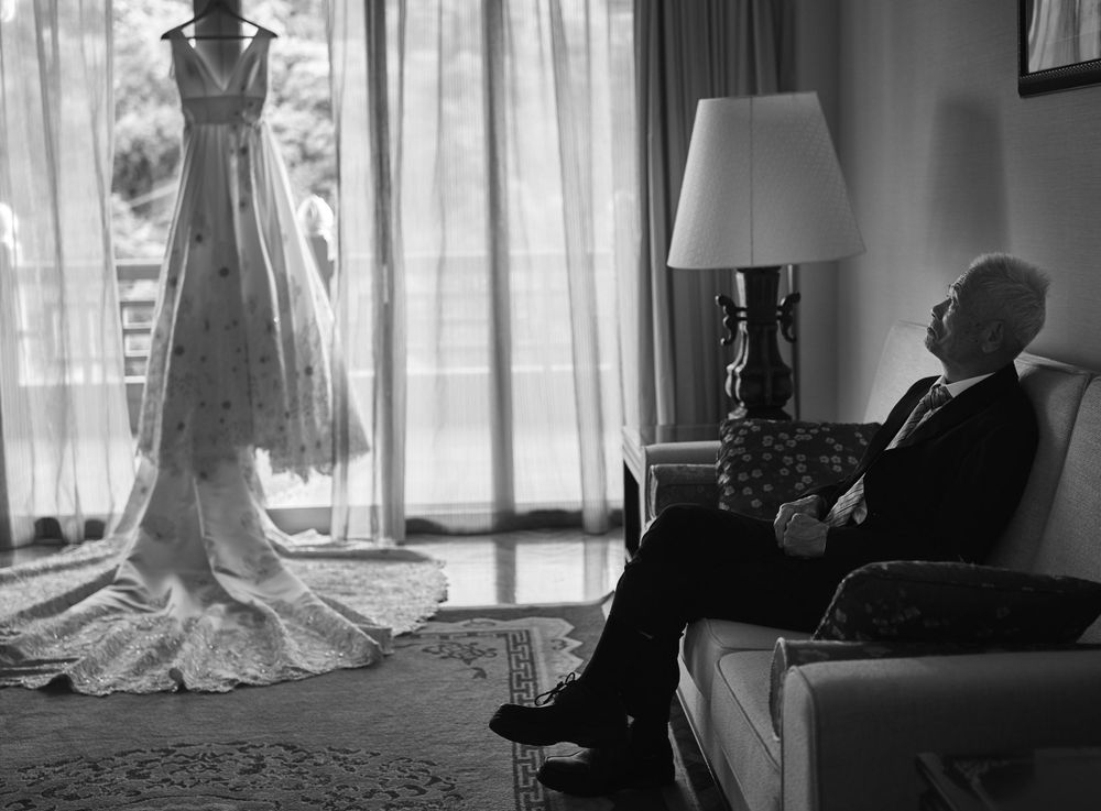 Elegant wedding dress hangs by window as a father sits in armchair in black and white vintage photograph.