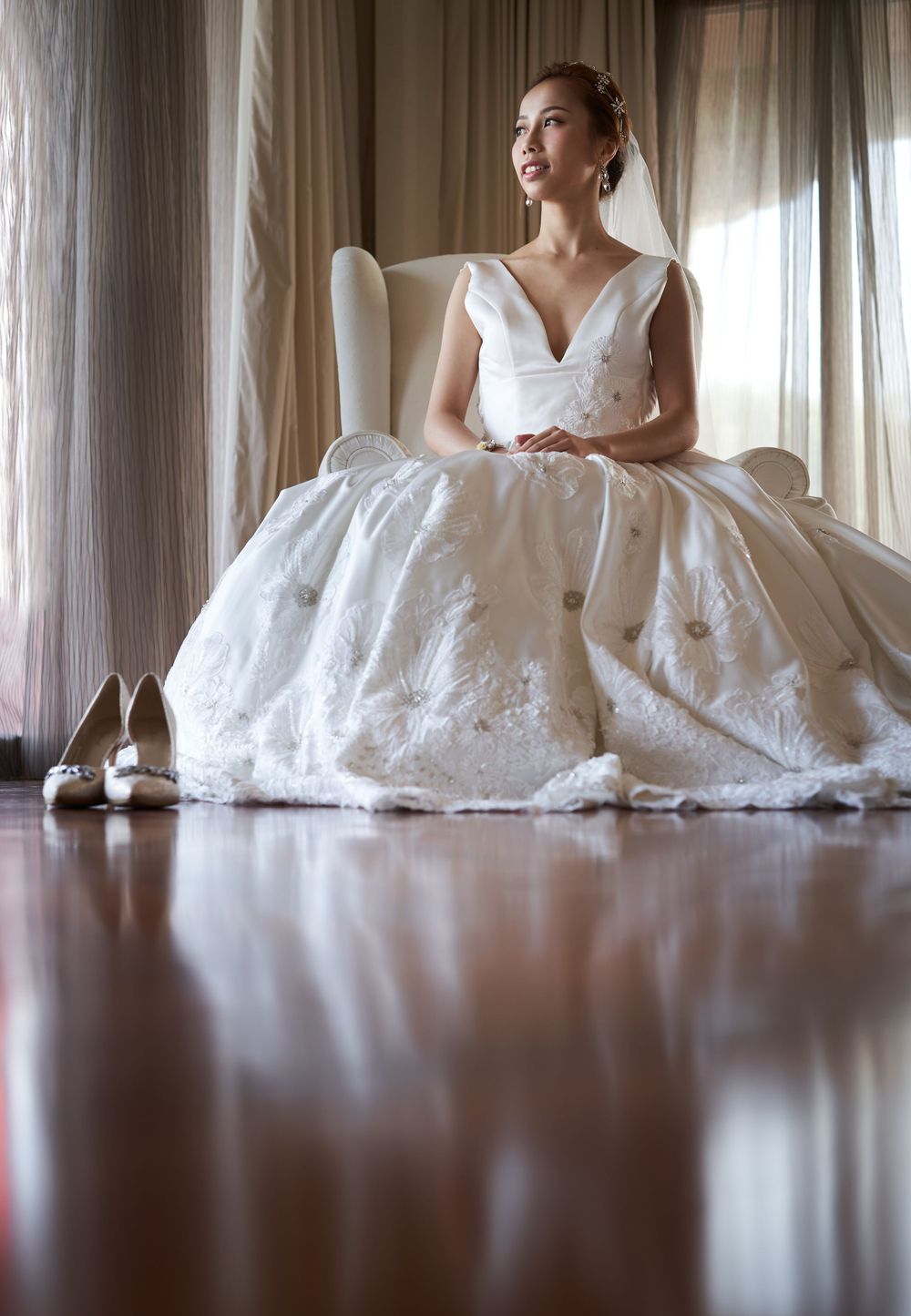 A bride in an elegant white ballgown sits on a wingback chair with wedding shoes visible on hardwood floor.