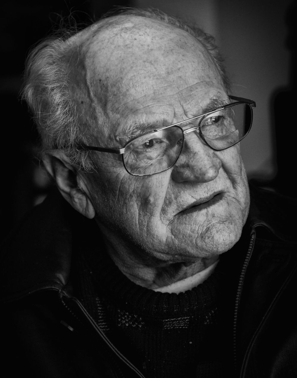 Black and white portrait of an elderly person wearing glasses and a dark sweater with a contemplative expression photographed by Evan Will
