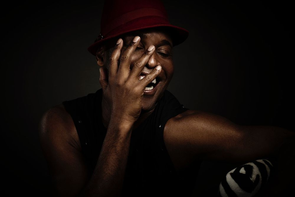 A dramatic portrait of a person wearing a red hat in dark lighting with their hand partially covering their face photographed by Evan Will