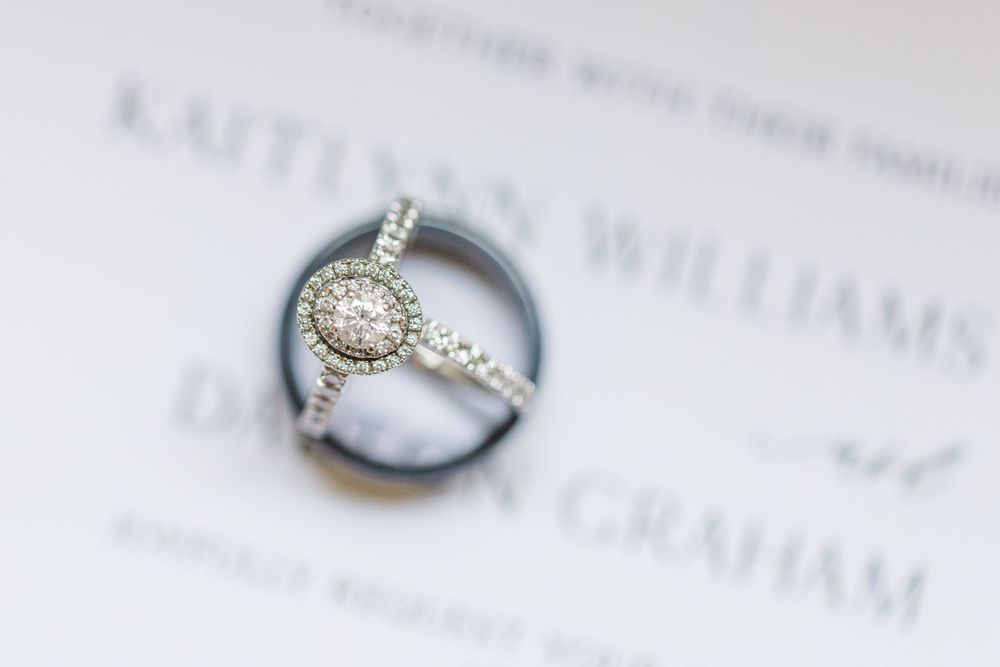 Elegant diamond engagement ring with halo design photographed against blurred text on white paper background.