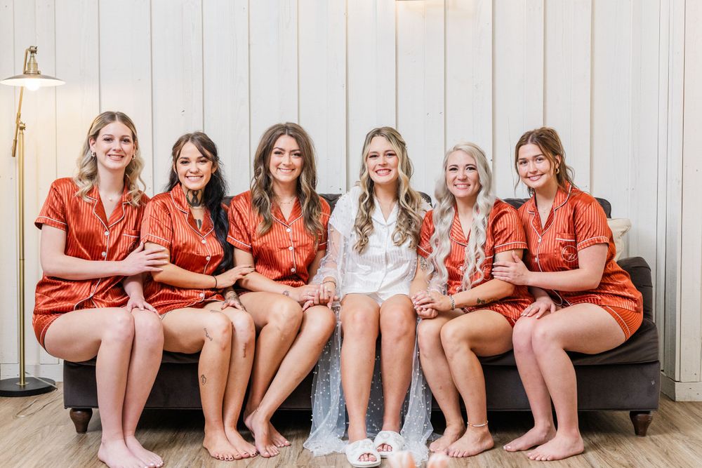 Wedding party in matching silk robes sits together on gray bench against white wall.