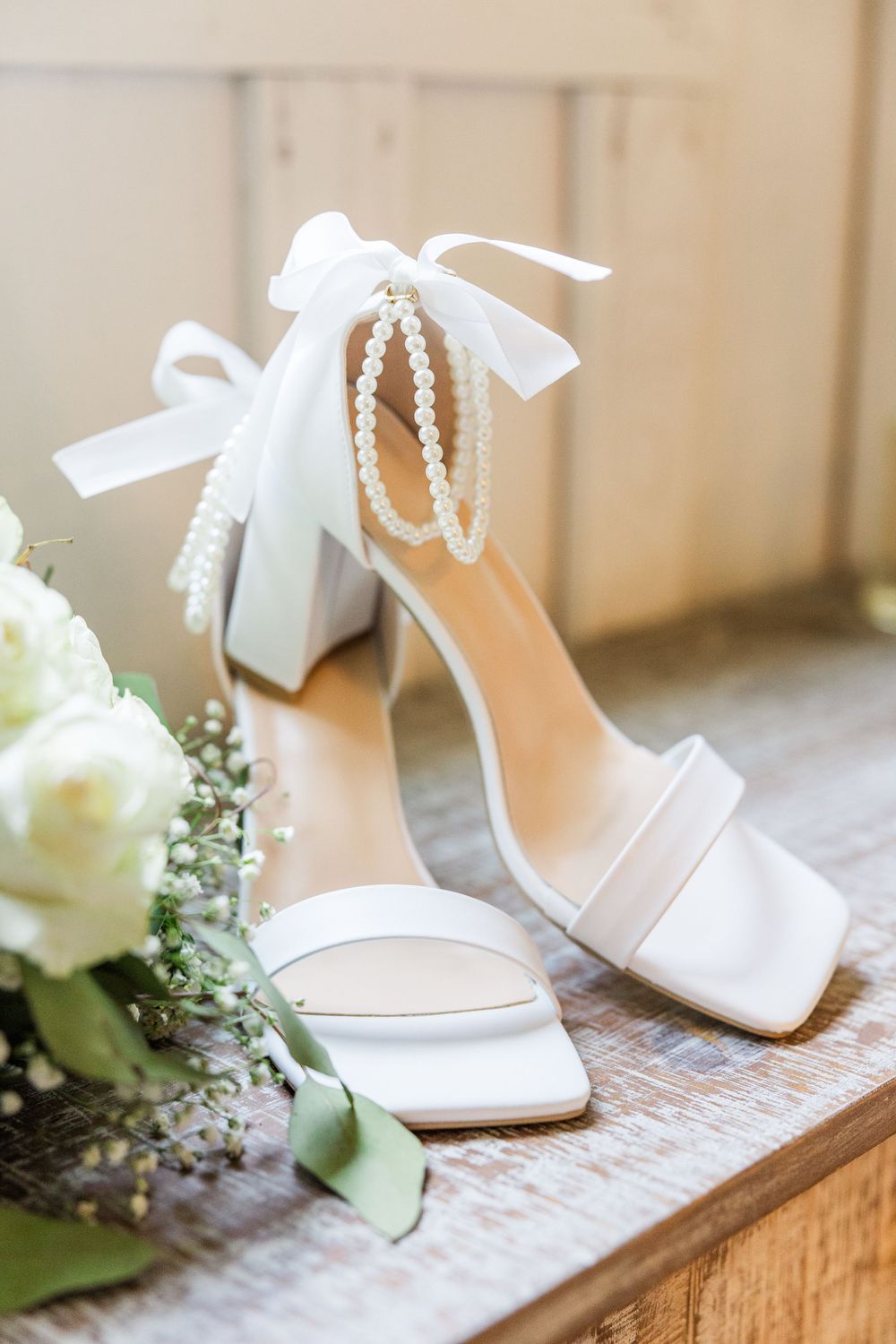 Elegant white bridal shoes with pearl ankle straps and bow details sit beside white roses.