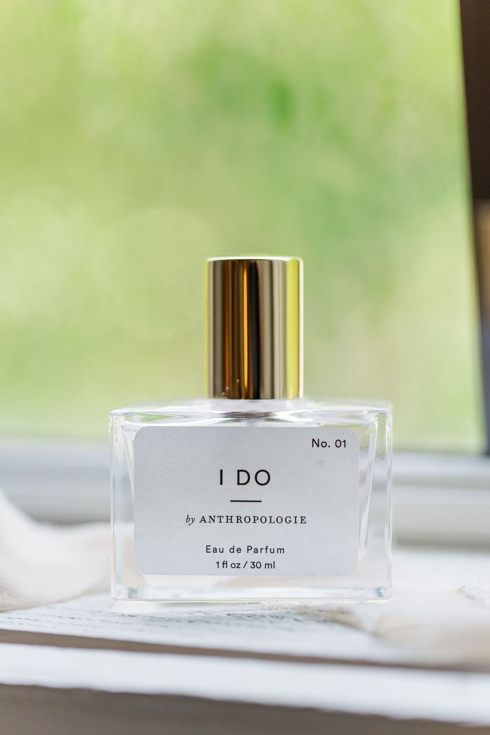 Glass perfume bottle labeled 'I DO' by Anthropologie displayed against a soft green blurred background.