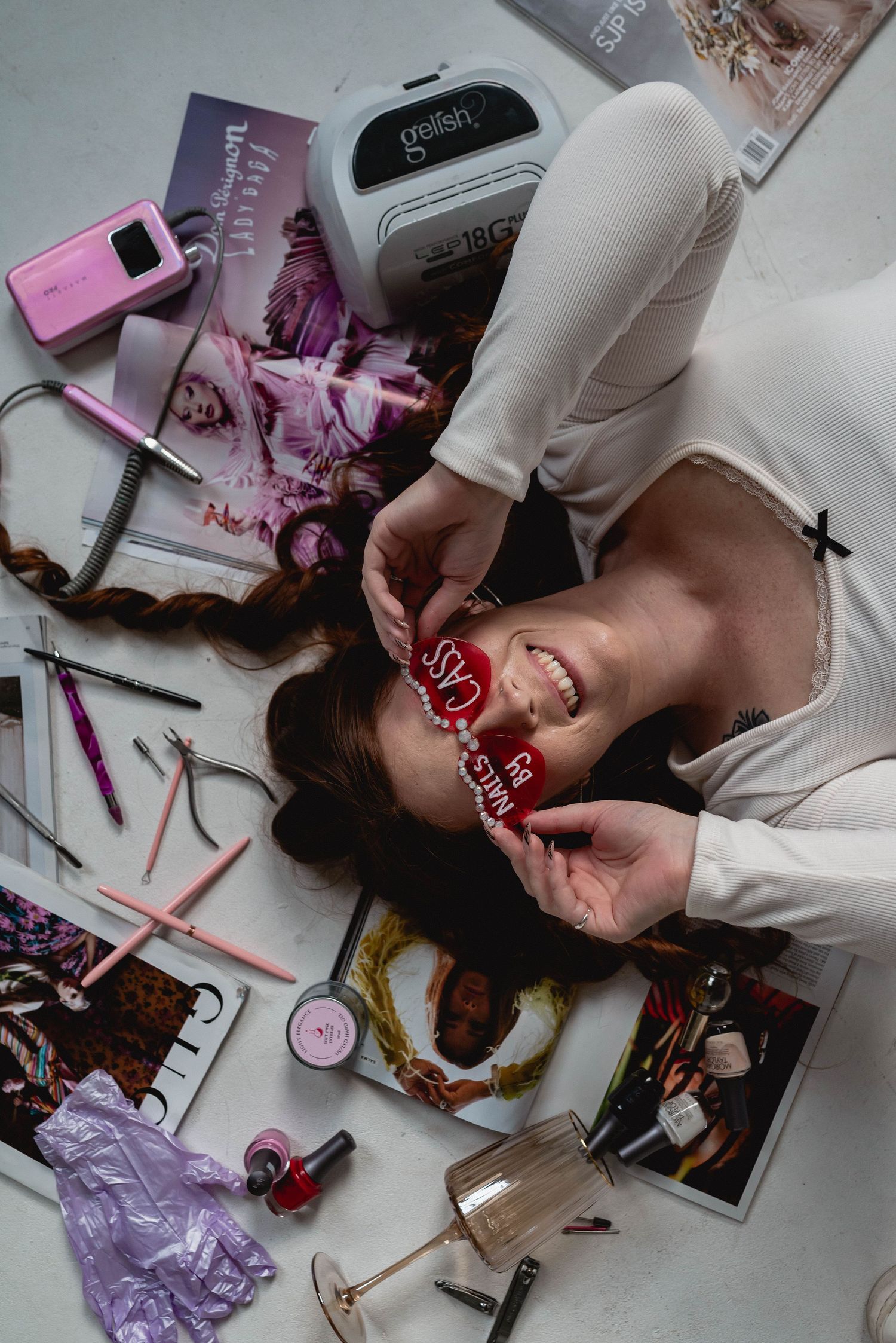 Flatlay of beauty products, magazines and accessories scattered on floor with someone wearing a white sweater.