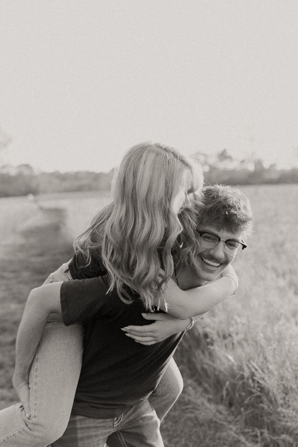 Black and white photo of a joyful piggyback ride in a grassy field during sunset, capturing a candid playful moment.