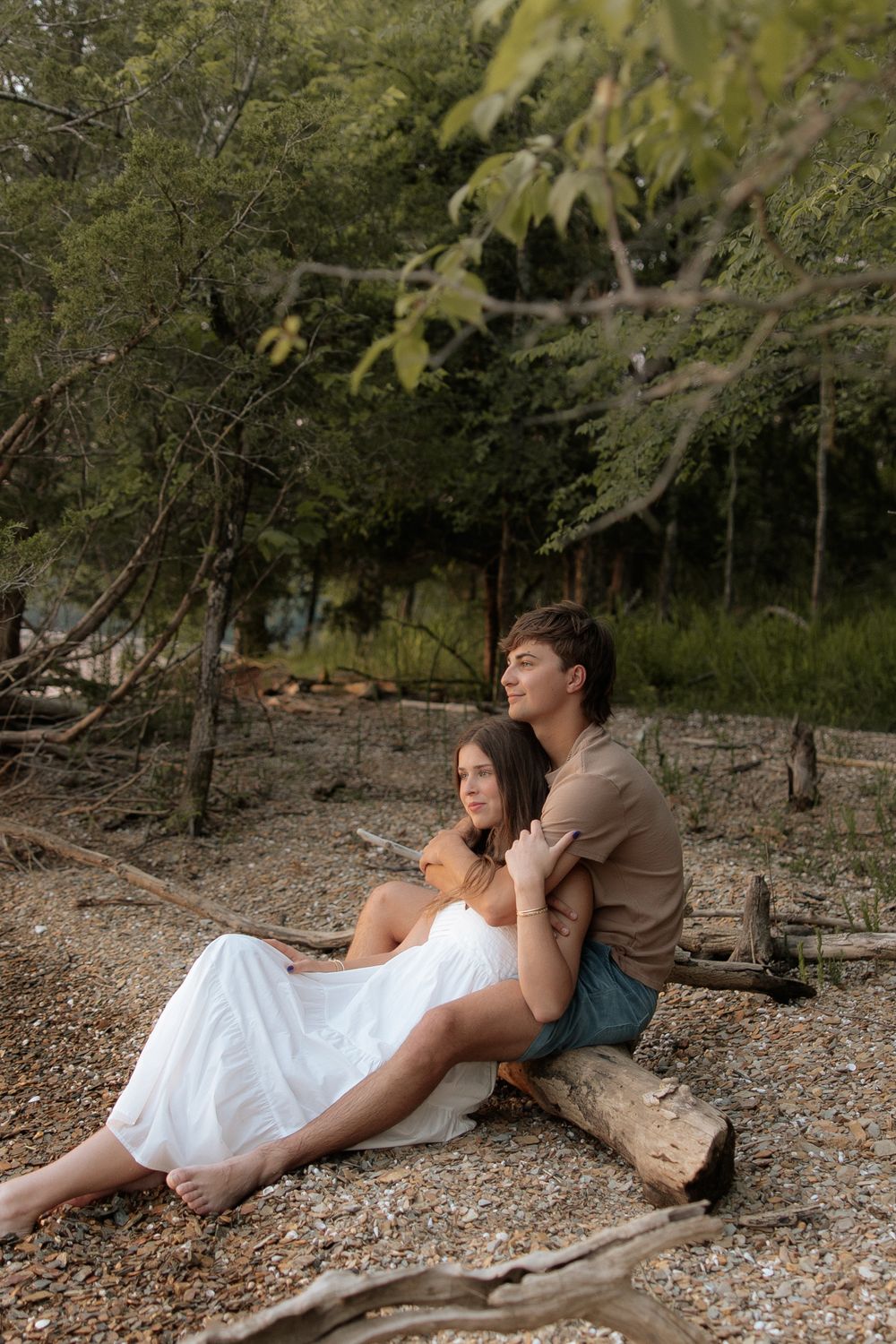 A couple embraces while sitting together on a log by the shore of a lake surrounded by trees.