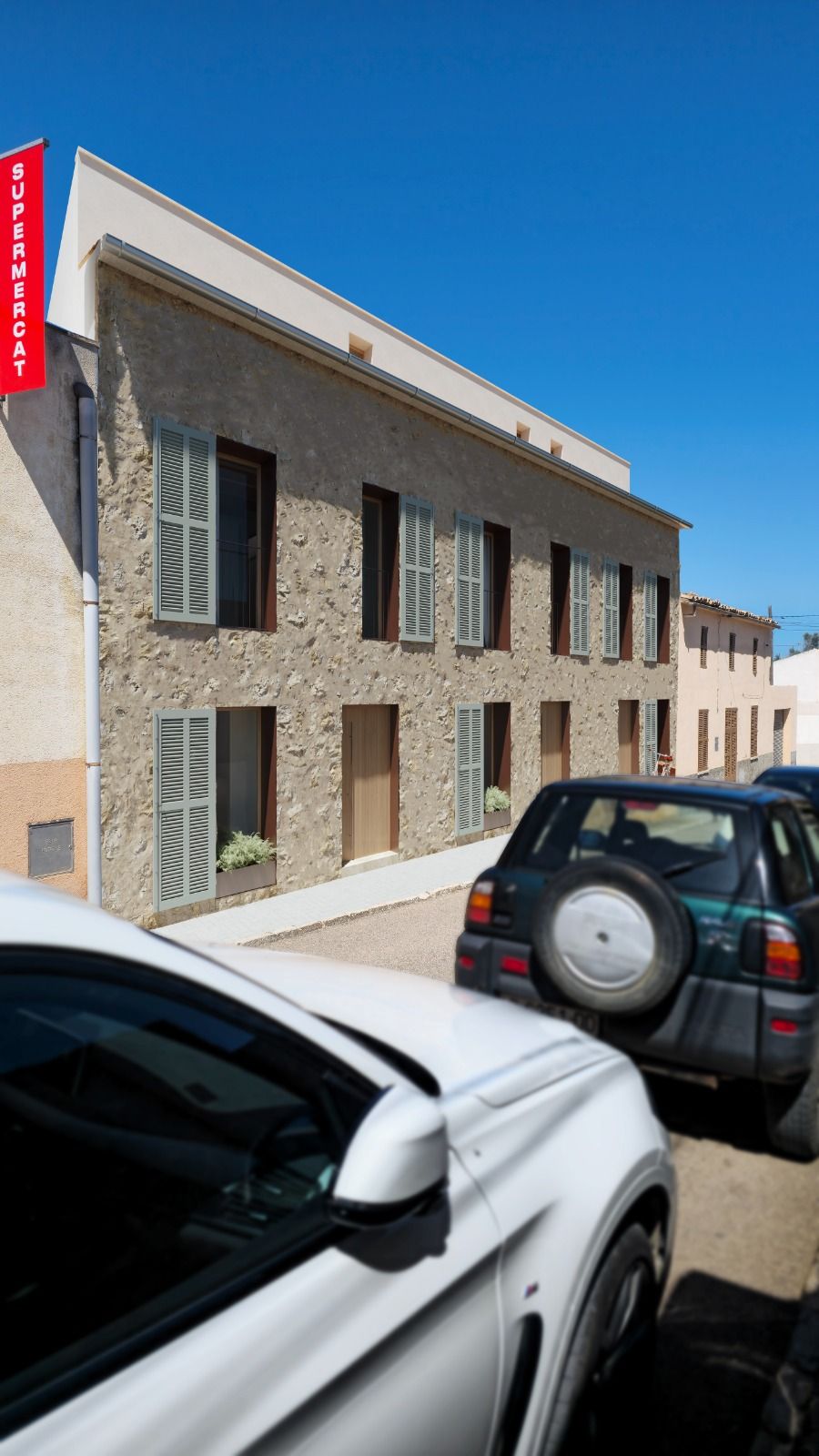 Two cars parked in front of a textured stucco building with shuttered windows and a red vertical sign on a sunny day.